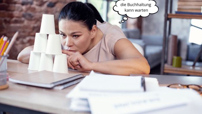 Gelangweilte Freelancerin baut Turm aus Pappbechern, Sprechblase: „die Buchhaltung kann warten“.