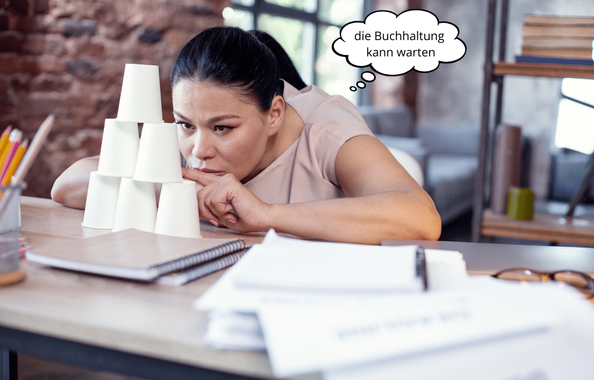 Gelangweilte Freelancerin baut Turm aus Pappbechern, Sprechblase: „die Buchhaltung kann warten“.